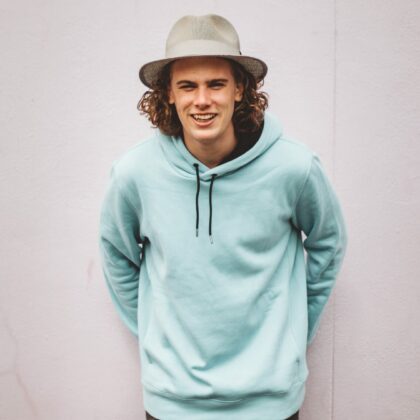 A young man wearing a blank hoodie and panama hat is leaning against a white wall. Custom custom clothing will provide full color custom hoodies for your business or event.