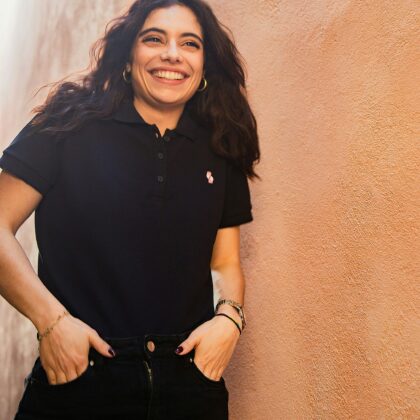 A woman wears a black customizable polo shirt and leans against a brown stucco wall. Custom custom clothing provides custom apparel services including custom embroidered polos.