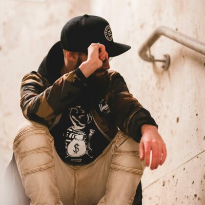 A young man sits in a concrete stairwell while wearing a ball cap. His head is tilted slightly to the left and his right hand is on the brim of his cap. Custom custom clothing creates custom apparel including various types of hats and caps.